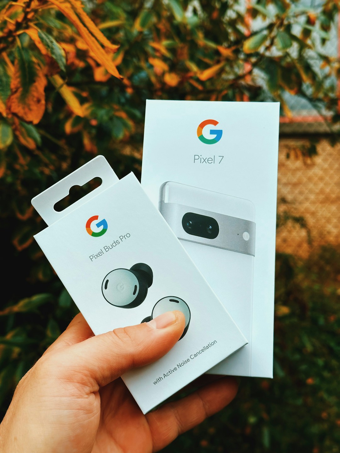 Google Pixel 7 and Pixel Buds Pro retail boxes held outdoors, showing Google's Pixel hardware lineup