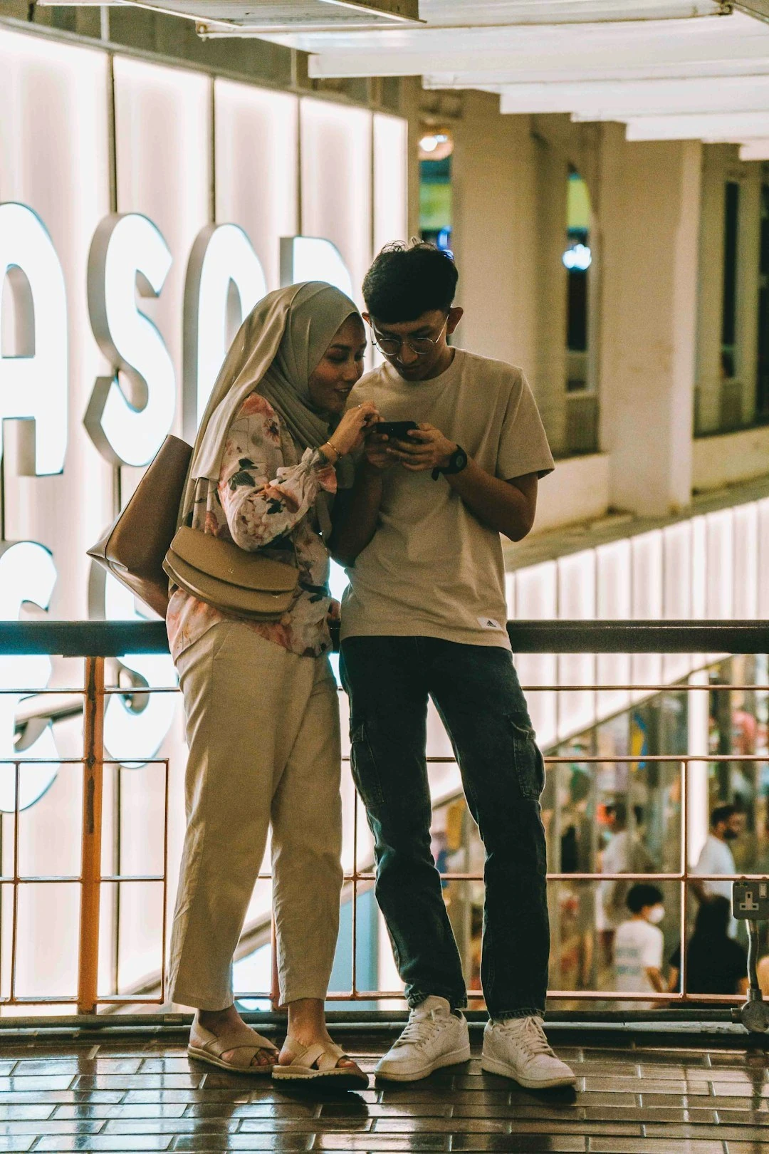 A couple checks a smartphone together at a brightly lit Malaysian shopping mall at night