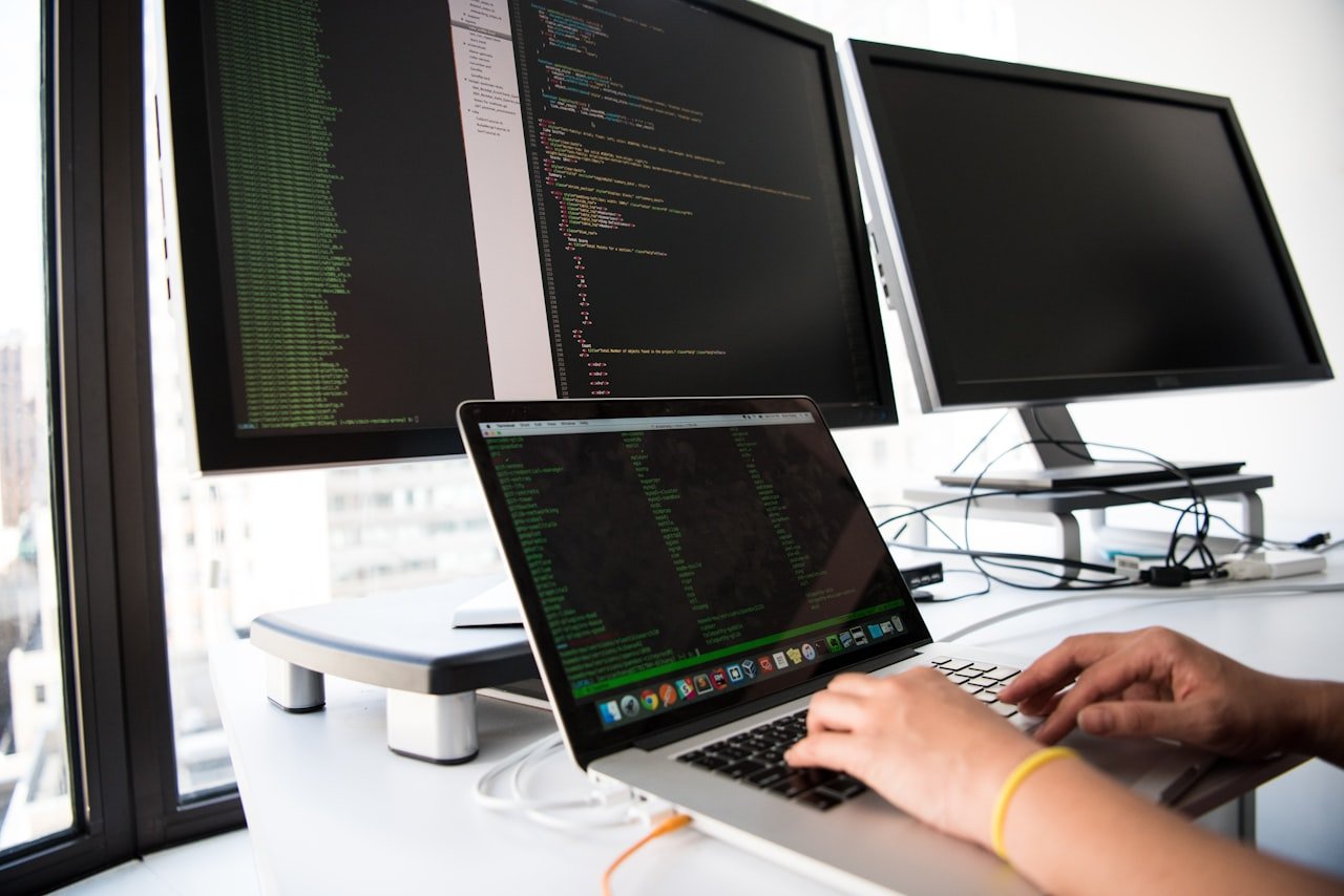 A developer typing on a laptop beside monitors filled with code, illustrating the AI tools businesses are racing to deploy