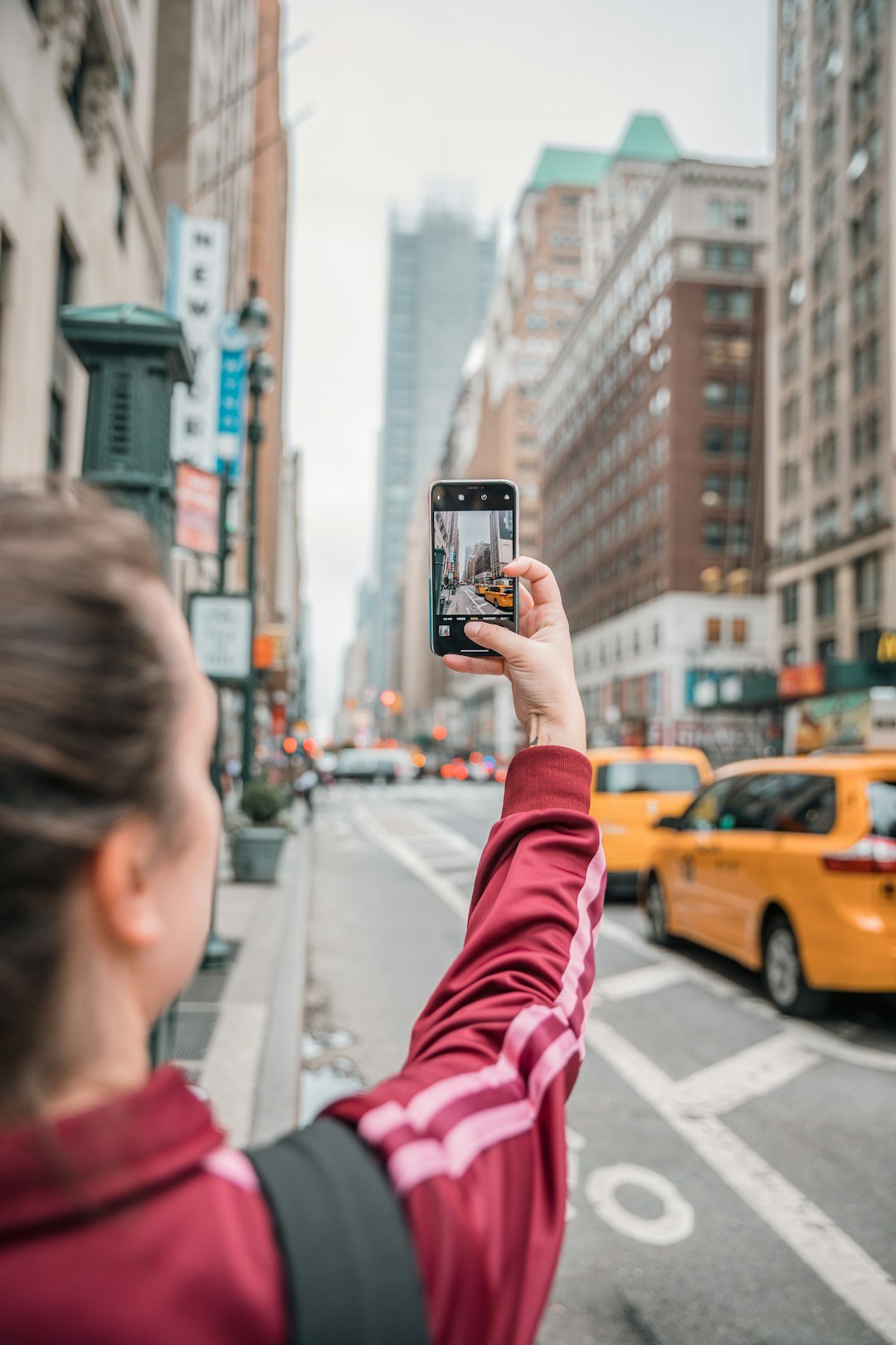 A person raising a smartphone to photograph a busy city street lined with tall buildings and yellow taxis
