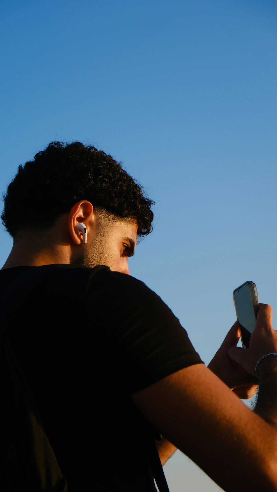 Seorang lelaki muda memakai AirPod putih di satu telinga sambil melihat telefon pintarnya di luar rumah