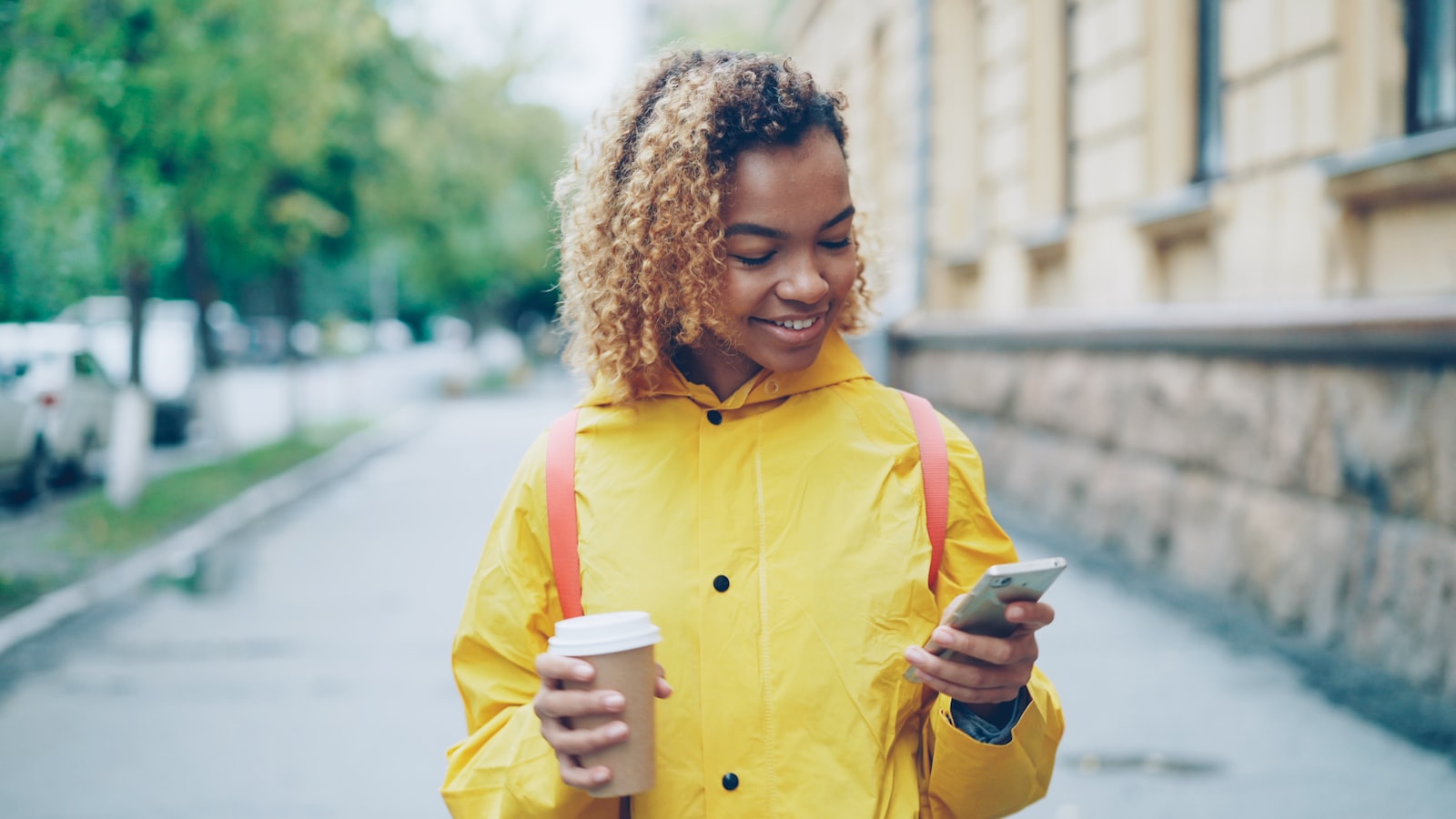 A person walks a city street looking at a smartphone with a takeaway coffee